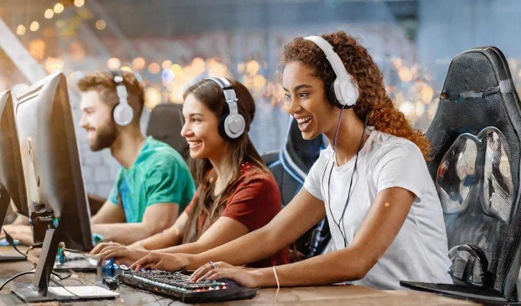 Group of three gamers wearing headphones and playing on computer stations, smiling and engaged.